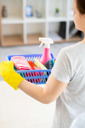 Cleaning service. Housework company. Home hygiene. Unrecognizable woman in protective gloves holding basket with cleaner spray soapy sponges brush microfiber cloth light room back view.の写真素材