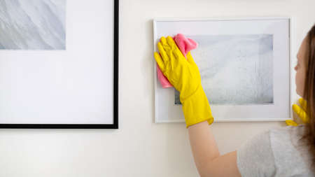 Home cleaning. Janitor woman. Housework routine. Unrecognizable woman in yellow protective gloves clearing picture art from dust microfiber cloth.の写真素材