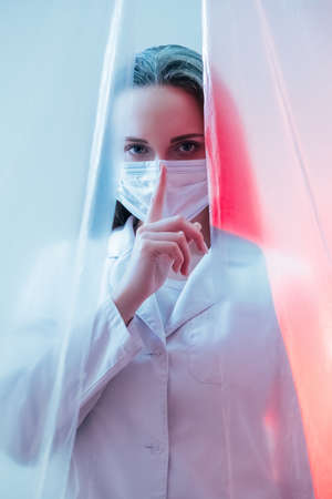 Medical silence. Hospital privacy. Pandemic quarantine. Female nurse in white coat face mask warning with hush finger gesture in pink blue neon light isolated behind plastic drapes.の写真素材