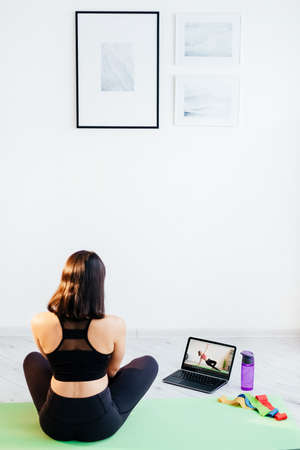 Fitness blog. Sportive woman. Home training. Lockdown lifestyle. Athletic lady in sportswear sitting on exercise mat with bottle of water stretching bands looking laptop with plank gym video.の写真素材