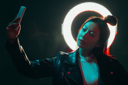Gadget people. Cyberpunk portrait. Futuristic technology. Blue red neon color light Asian girl in black leather jacket taking selfie on phone with white halo in smoke at dark night.の写真素材