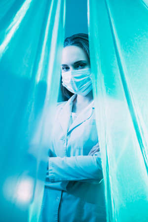 Frontline doctor. Medic portrait. Healthcare professional. Female physician in white coat protective face mask standing crossed arms isolated behind blue plastic drapes.の写真素材