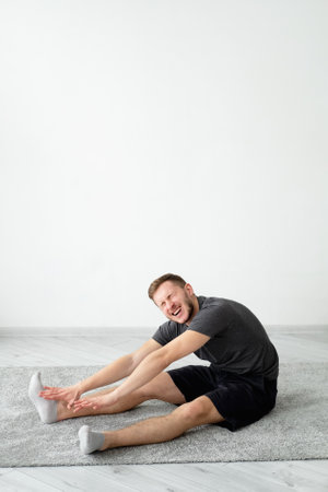 Positive man. Home relax. Lockdown lifestyle. Harmony peace. Handsome happy guy in t-shirt shorts sitting floor carpet in lotus pose leaning hand on leg light wall interior background copy space.の写真素材