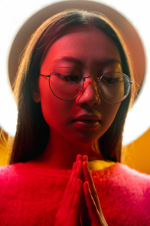 Cyberpunk people. Spiritual portrait. Future faith. Blue neon light art portrait of Asian prayer girl in black leather jacket namaste with white halo in red color smoke on dark.の写真素材