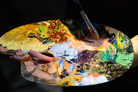 Color life. Creative process. Art tools. Painting artwork. Unrecognizable woman holding palette with blending colorful paints and brush isolated dark.の写真素材