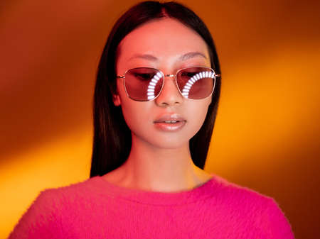 Color light face. Fashion portrait. Eyewear accessories. Pink neon confident glamour Chinese girl in stylish sunglasses isolated on bright yellow golden gradient background.の写真素材