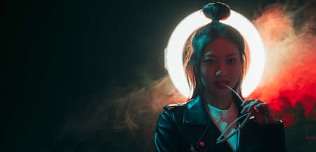 Night fashion banner. Cyberpunk people. Futuristic beauty. Stylish Asian girl in black leather jacket with white halo ring in red neon color light mist on dark empty space background.の写真素材