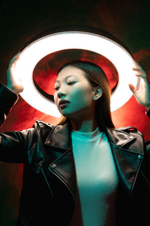 Cyberpunk fashion. Futuristic beauty. Sci-Fi technology. Blue neon light art portrait of Asian girl in black leather jacket with white glowing halo in red color smoke at dark night.の写真素材