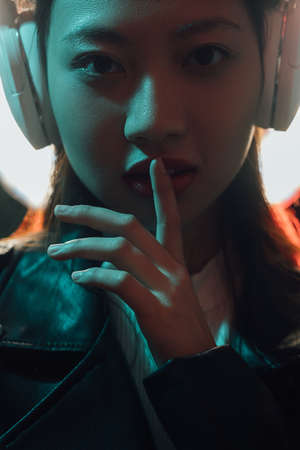 Neon light face. Cyber music. Urban subculture. Futuristic mystery. Night blue red color portrait of Asian girl in headphones showing hush gesture on dark glowing background.の写真素材