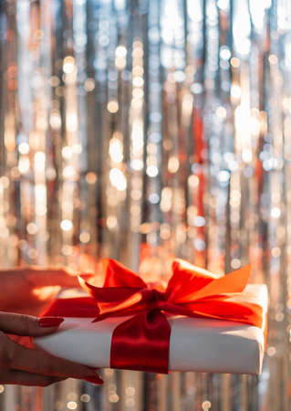 Birthday present. Holiday decoration. Greeting congratulation. Female hands holding gift box red ribbon on shimmering sparkle silver cascade curtain blur lights.の写真素材