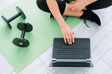 Quarantine sport. Online training. Weight fitness. Home gym. Strong body. Unrecognizable woman sitting lotus pose scrolling laptop dumbbells near on mat in light room interior.の写真素材