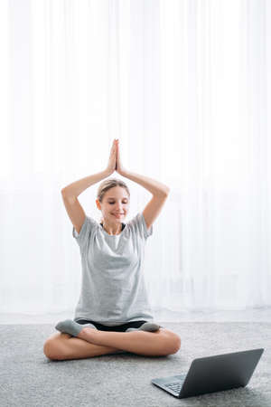 Online yoga. Home training. Morning relaxation. Peaceful girl sitting cross-legged on floor watching video tutorial on laptop on light white window background.の写真素材