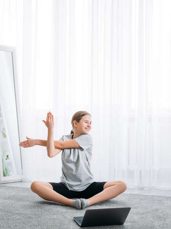 Fitness online. Home training. Active kids. Physical health. Girl doing stretching exercise on floor alone with laptop at light white window interior.の写真素材