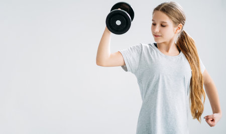 Kids sport. Fitness equipment. Gym workout. Physical exercise. Strong athletic girl in training arm muscles with dumbbell isolated on light copy space background.の写真素材