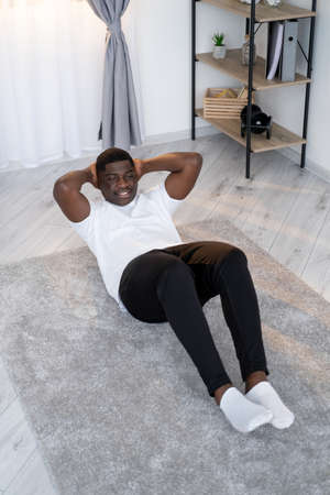 Abs training. Sportive black man. Home fitness. Healthy body. Enjoying sport. Happy african guy doing trunk curls workout on floor carpet in light room interior.の写真素材
