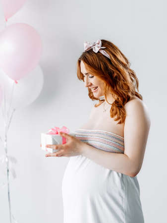 Baby shower party. Pregnant woman. Festive surprise. Happy motherhood. Smiling tummy lady holding gift box pink ribbon isolated white pastel balloons decoration.の写真素材