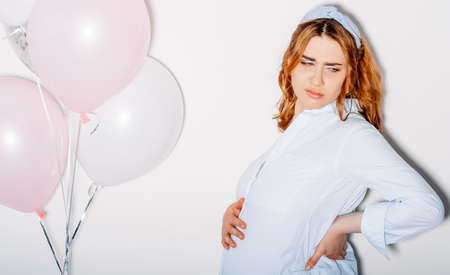 Hard pregnancy. Tired woman. Maternity feelings. Child expectation problems. Gloomy tummy lady looking upset touching baby bump and back isolated white light balloons decor copy space.の写真素材