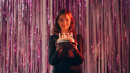 Birthday cake. Happy woman. Holiday celebration. Pretty smiling lady elegant dress holding festive pastry with candle lights on shimmering purple cascade curtain background.の写真素材
