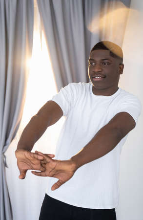 Morning exercise. Happy black man. Enjoying sport. Healthy lifestyle. Smiling african guy white t-shirt stretching hands sunshine lens flare light room interior.の写真素材