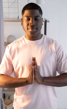 Morning relax. Peaceful black man. Home yoga. Harmony mind. Smiling african guy meditating holding namaste hands in sunshine light room interior.の写真素材