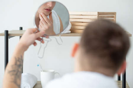 Morning hygiene. Facial treatment. Skincare procedure. Grooming routine. Young handsome man touching soft clean perfect face in mirror reflection in light bathroom interior.の写真素材