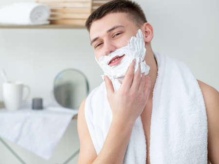 Hygiene morning. Cosmetic product advertising. Male grooming. Joyful smiling young shirtless guy white towel on shoulders applying shaving cream on face in light bathroom.の写真素材
