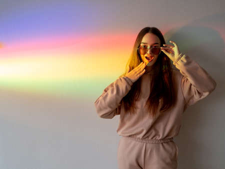 Surprised girl. Fashion background. Discount sale. Shock emotion. Attractive impressed female with omg gesture wearing sunglasses on pastel rainbow ray color neon lights copy space background.の写真素材