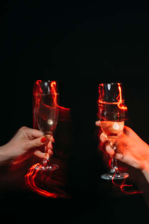 Corporate party. Holiday toast. Cheer greeting. Festive celebration. Female hands holding glasses of champagne long exposure defocused red light isolated black.の写真素材
