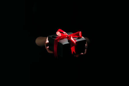 Christmas present. Festive surprise. Stylish decor. Unrecognizable woman holding elegant black gift box red ribbon in hands isolated dark light glow defocused.の写真素材