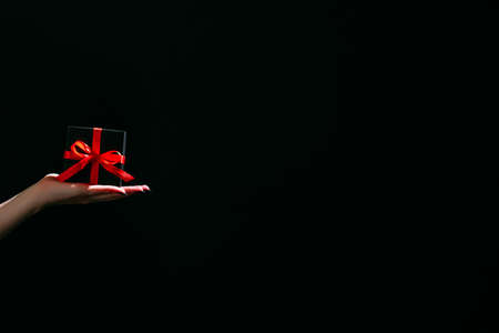 Christmas gift. Festive surprise. Holiday congrats. Advertising background. Unrecognizable woman holding elegant black gift box red ribbon in hands isolated dark copy space.の写真素材