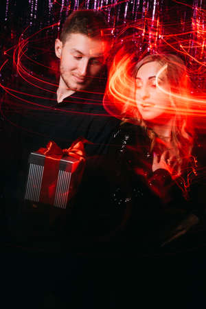 Couple congratulation. Christmas surprise. Happy New Year 2022. Festive night greeting. Woman impressed by boyfriend gift box in blur red neon light strokes effect on dark tinsel.の写真素材