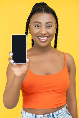 Mobile application. Phone advertising. Online service. Happy smiling Afro woman demonstrating smartphone mockup isolated on orange background.の写真素材
