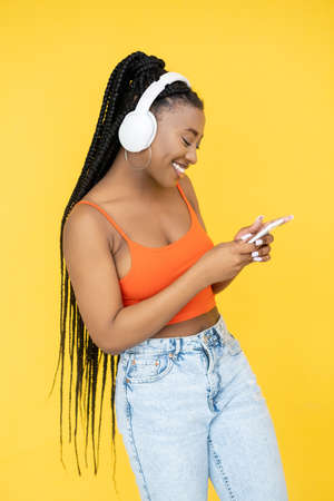 Music gadget. Millennial lifestyle. Mobile technology. Happy African woman in headphones listening playlist on phone isolated on yellow background.の写真素材