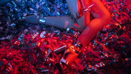 Drunk woman. Christmas celebration. Night club party. Unrecognizable lady elegant golden shoes sitting floor with champagne glass on holiday confetti ribbon decor blue red neon light.の写真素材