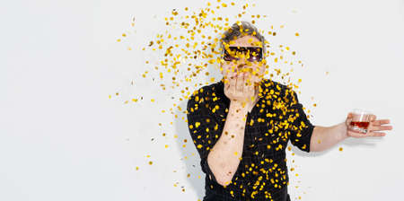 Holiday banner. Party time. Festive celebration. New Year. Happy man in sunglasses with alcohol glass blowing golden confetti isolated on white copy space studio background.の写真素材