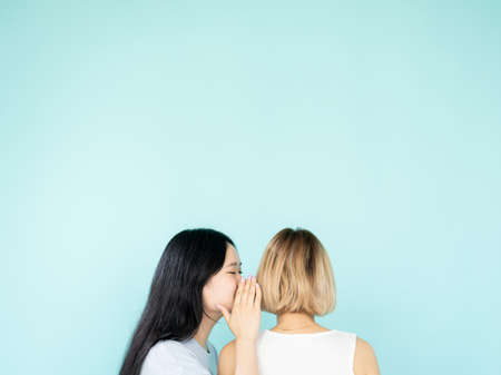 Sharing secrets. Conspirator women. Gossip spreading. Unrecognizable asian lady whispering information to blonde friend isolated blue copy space.の写真素材