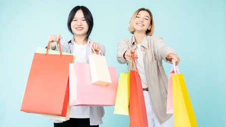 Shopaholic lifestyle. Happy women. Hot sale. Best offer. Shopping discount. Pretty smiling asian caucasian friends showing colorful purchase bags isolated blue.の写真素材