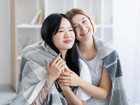 Enjoying meeting. Ethnic friendship. Happy women. Pretty smiling caucasian asian ladies embracing covering plaid light room interior.の写真素材