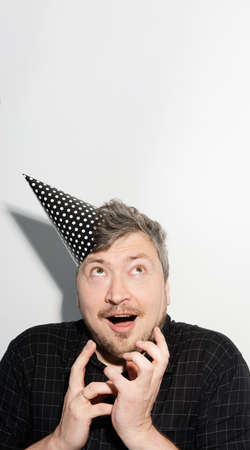 Birthday surprise. Party celebration. Holiday fun. Festive card. Excited amazed happy smiling man in cone hat looking up at empty space wish greeting white studio background.の写真素材