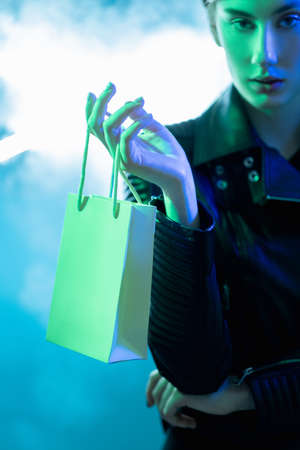 Shopping sale. Cyber monday. Black Friday. Special offer. Blue green neon color light confident woman in leather jacket holding bag on smoke cloud background.の写真素材