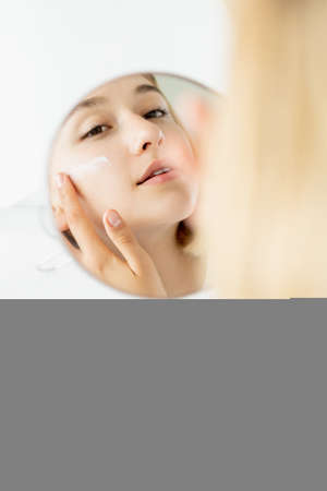 Facial care. Moisturizing product. Morning treatment. Pretty woman applying anti-aging cream on smooth face at mirror in white free space.の写真素材