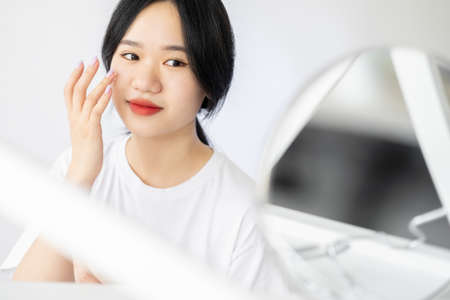 Morning facial care. Asian woman. Moisturizing treatment. Skincare cosmetic. Pretty lady applying face cream looking mirror in light room interior copy space.の写真素材