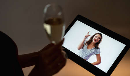Online birthday. Festive greeting. Virtual party. Unrecognizable woman with glass of champagne looking happy female friend congratulating on tablet computer in dark room interior.の写真素材