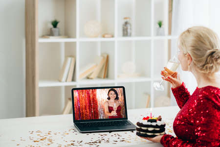 Christmas celebration. Virtual greeting. Distance party. Festive woman drinking champagne holding chocolate cake looking female friend toast on laptop in light room interior.の写真素材