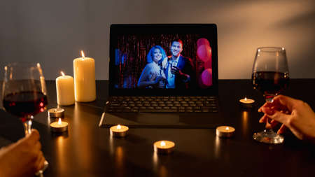 Romantic meeting. Virtual celebration. Anniversary party. Unrecognizable couple glass wine celebrating with friends on laptop in festive candles decorated room interior.の写真素材