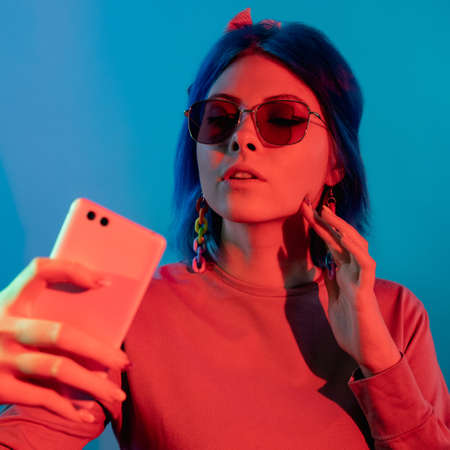 Neon light selfie. Futuristic technology. Digital people. Glamorous millennial woman taking photo on cell on red color shine isolated on blue background.の写真素材