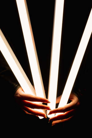 Light tube. LED illumination. Modern lighting. Closeup of female hands holding bright glowing fluorescent lamp isolated on black background.の写真素材