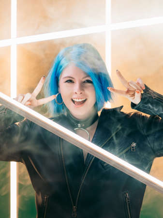 Peace gesture. Cyberpunk party. Synthwave style. Excited smiling woman showing victory hand sign on LED lamp light on smoke background.の写真素材