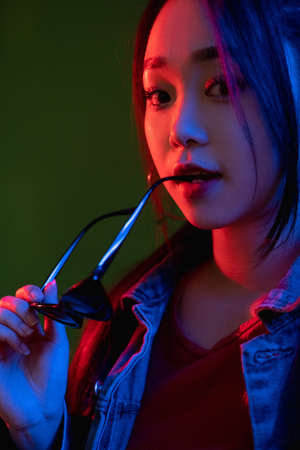 Neon light portrait. Futuristic model. Synthwave style. Gorgeous woman face biting sunglasses in...