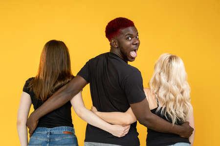Open relationship. Casanova man. Multiracial love. Date luck. Lovelace successful amazed happy African guy embracing two different women together isolated on orange background.の写真素材